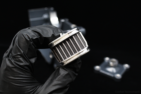 A gloved hand holds a square Conflux heat exchange cartridge with gear box housing in background